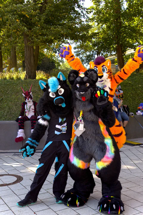 Photo of a black and blue wusky fursuiter and me in black jaguar fursuit, waving at the camera, while a bright orange tiger fursuiter is jumping behind us, paws in the air