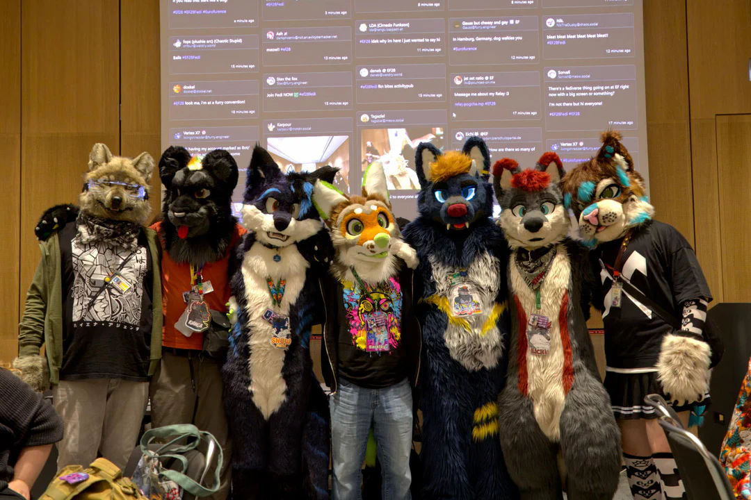 Photo of seven fursuiters posing in front of a wall with projected Fediverse posts