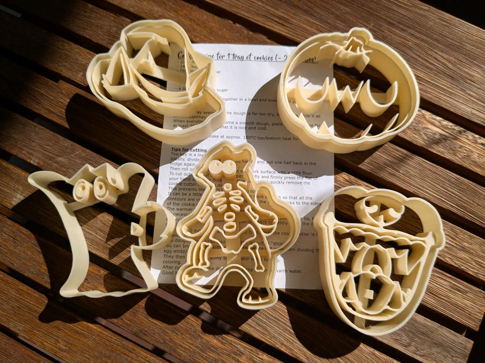 Photo of five furry Halloween themed cookie cutters in various shapes: a wizard hat with ears poking through, a pumpkin with the text “UWU”, a ghostly cat, an anthropomorphic feline skeleton, and something that looks like a banner with teeth?