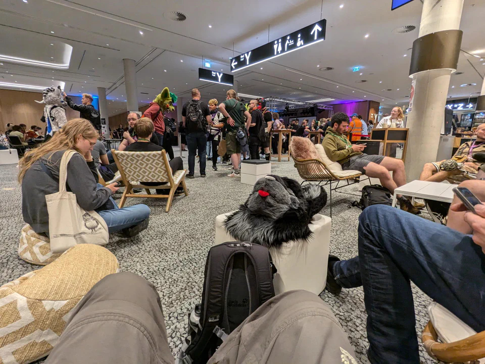 First-person photo of me sitting in the area near the open stage, my legs are visible, with my fursuit head and backpack next to me on a chair and lots of other people around me