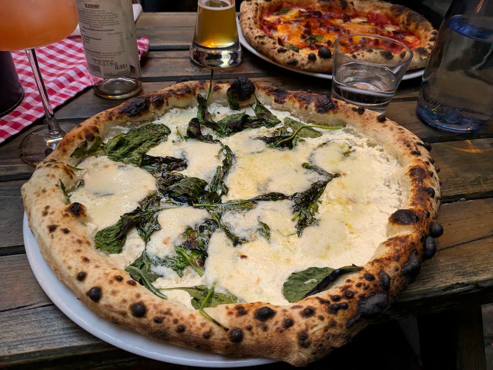 Photo of a pizza with spinach on a wooden table, next to colorful cocktails