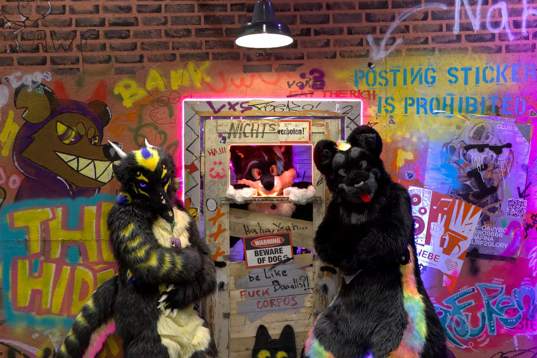 Photo of two fursuiters, a dragon and a black jaguar, standing in front of a door and wall covered in graffiti with arms crossed, looking like bouncers, while the third, an arctic fox, looks through a window in the door